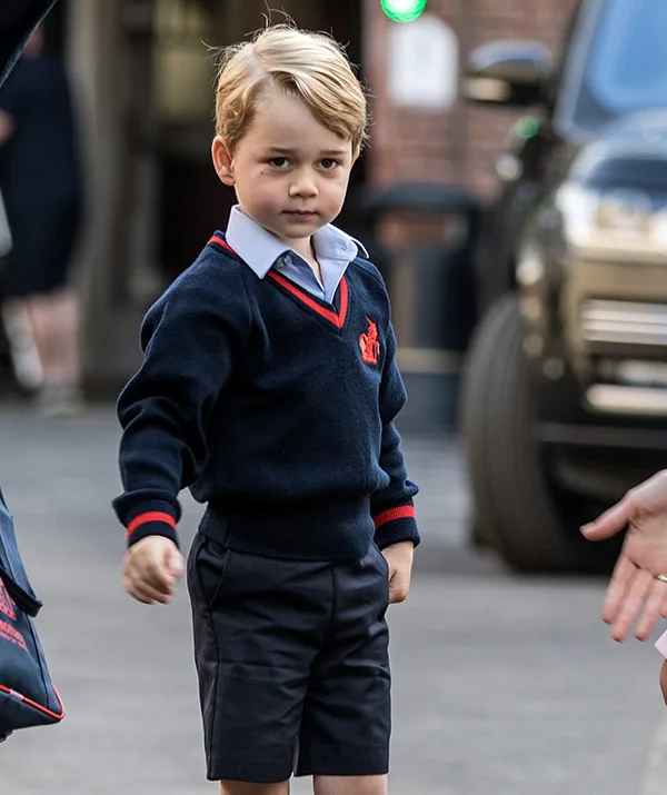 Young child in a navy school uniform standing outdoors, holding someone's hand.