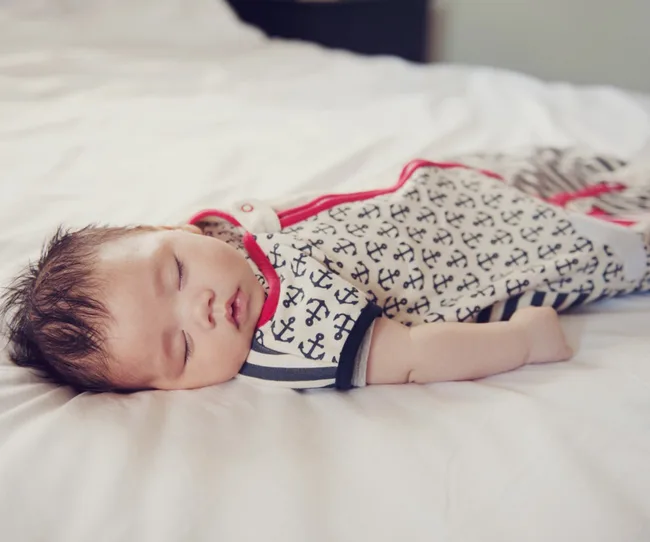 Sleeping baby in anchor-patterned onesie on a bed.