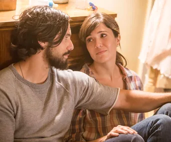 A man and woman sit closely on the floor, sharing an intimate and tender moment in a warmly lit room.