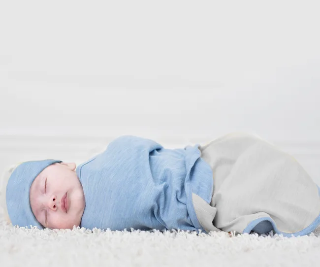 A sleeping baby wrapped in a blue swaddle lying on a soft white carpet.