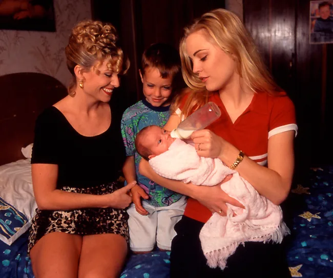Two women smiling at a baby being bottle-fed by a young woman, with a young boy watching nearby, in a cozy room setting.