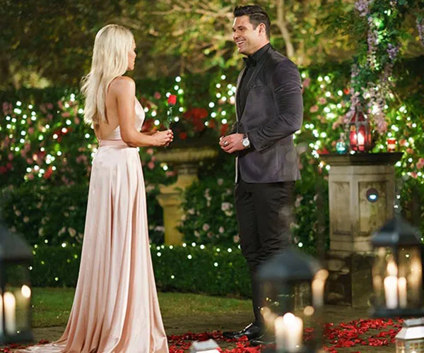 A woman in a flowing gown talks to a man in a suit at a romantically lit garden setting, surrounded by flowers and lanterns.