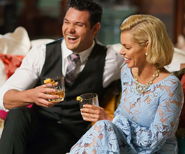 Man and woman in formal attire laughing and holding drinks on a couch.