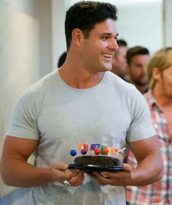 Man in a gray shirt holding a small cake with colorful candles, smiling, with people in the background.
