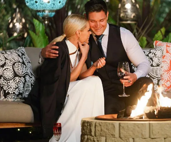 A couple sits close by an outdoor fire pit, holding wine glasses and smiling warmly at each other.