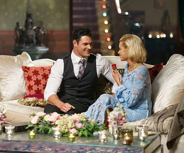 A couple sits on a sofa, smiling at each other, surrounded by flowers and candles, in a cozy, warmly lit room.
