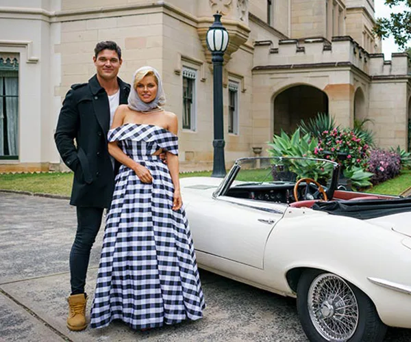 Man and woman standing by a white vintage convertible outside a large stone building, woman wearing a checkered dress.
