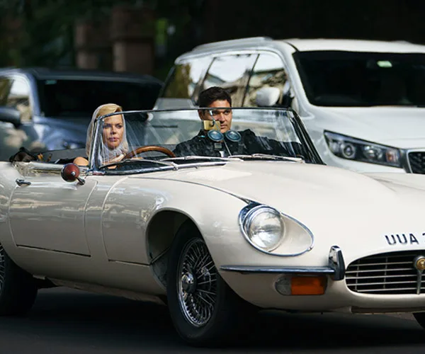 Convertible car with two people, a blonde woman and a man with sunglasses, driving through a street with other cars.