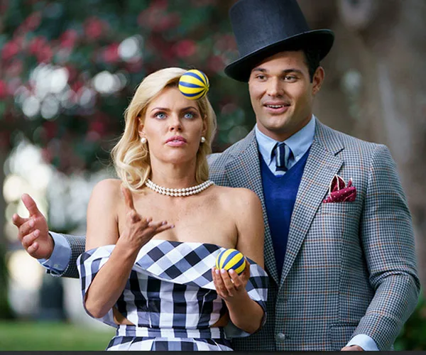 Man in a top hat assisting a woman in juggling two balls outdoors, both in stylish attire.