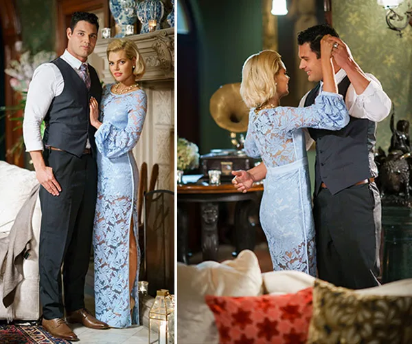 A couple elegantly dressed in a blue lace gown and vest poses indoors; woman adjusts man's collar in a cozy room.