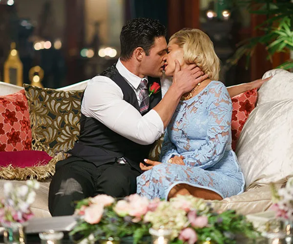 A couple kissing on a couch adorned with floral cushions in a dimly lit, elegant setting.