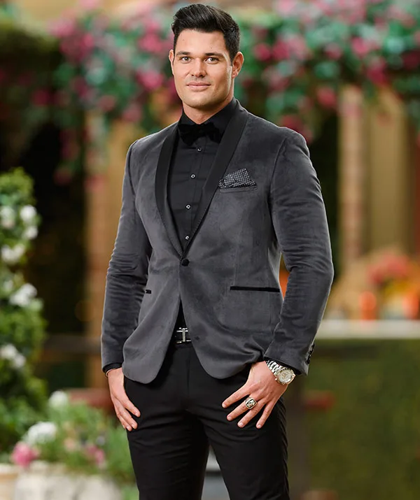 Man in a gray suit with black shirt and bow tie, standing outdoors with blurred floral background.