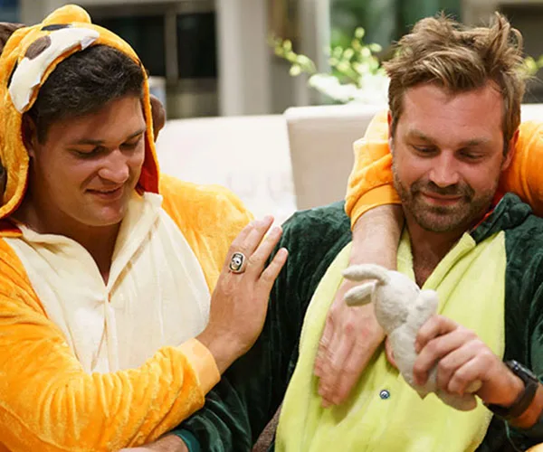 Two men dressed in animal onesies smiling and playing with a plush toy indoors.
