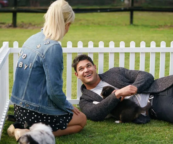 A man lies on grass inside a small fence, holding a puppy and smiling at a woman with blonde hair wearing a denim jacket.