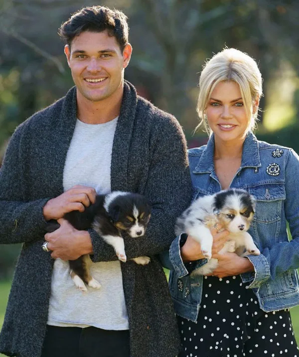 Two people smiling, each holding a puppy in a sunny outdoor setting.