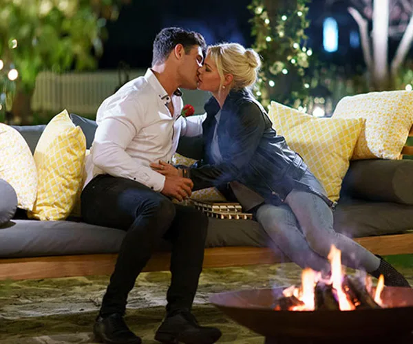 A couple kissing on an outdoor couch by a fire pit, surrounded by cushions, in a warm and romantic setting.