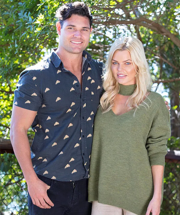 A man and woman standing outdoors, smiling, with trees in the background.