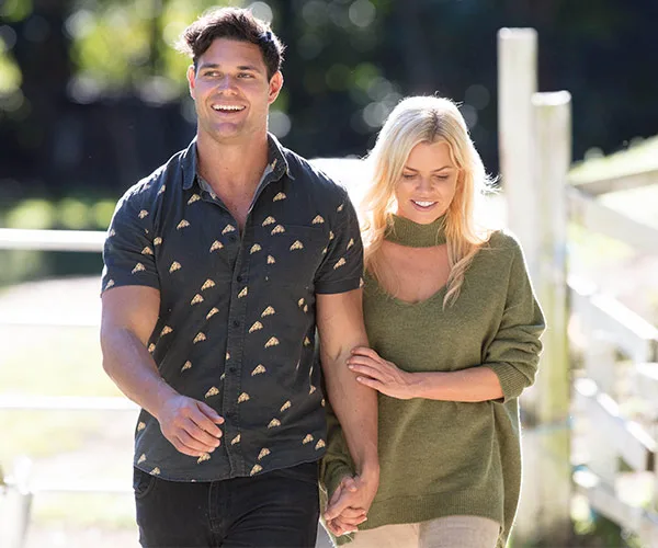 A couple walking outdoors, holding hands, with a sunny background and trees.