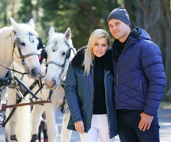 Couple in winter clothing stands next to two white horses in a forest setting.