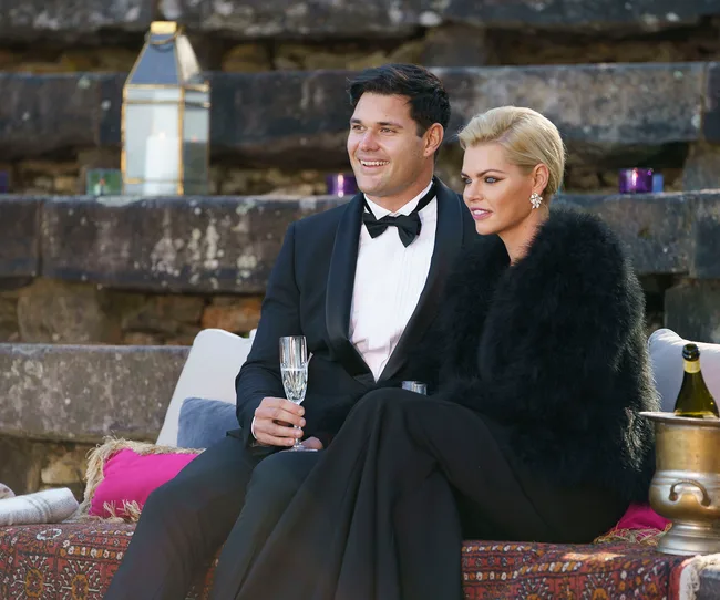 Couple dressed formally, sitting on a couch outdoors with champagne, stone steps in the background.