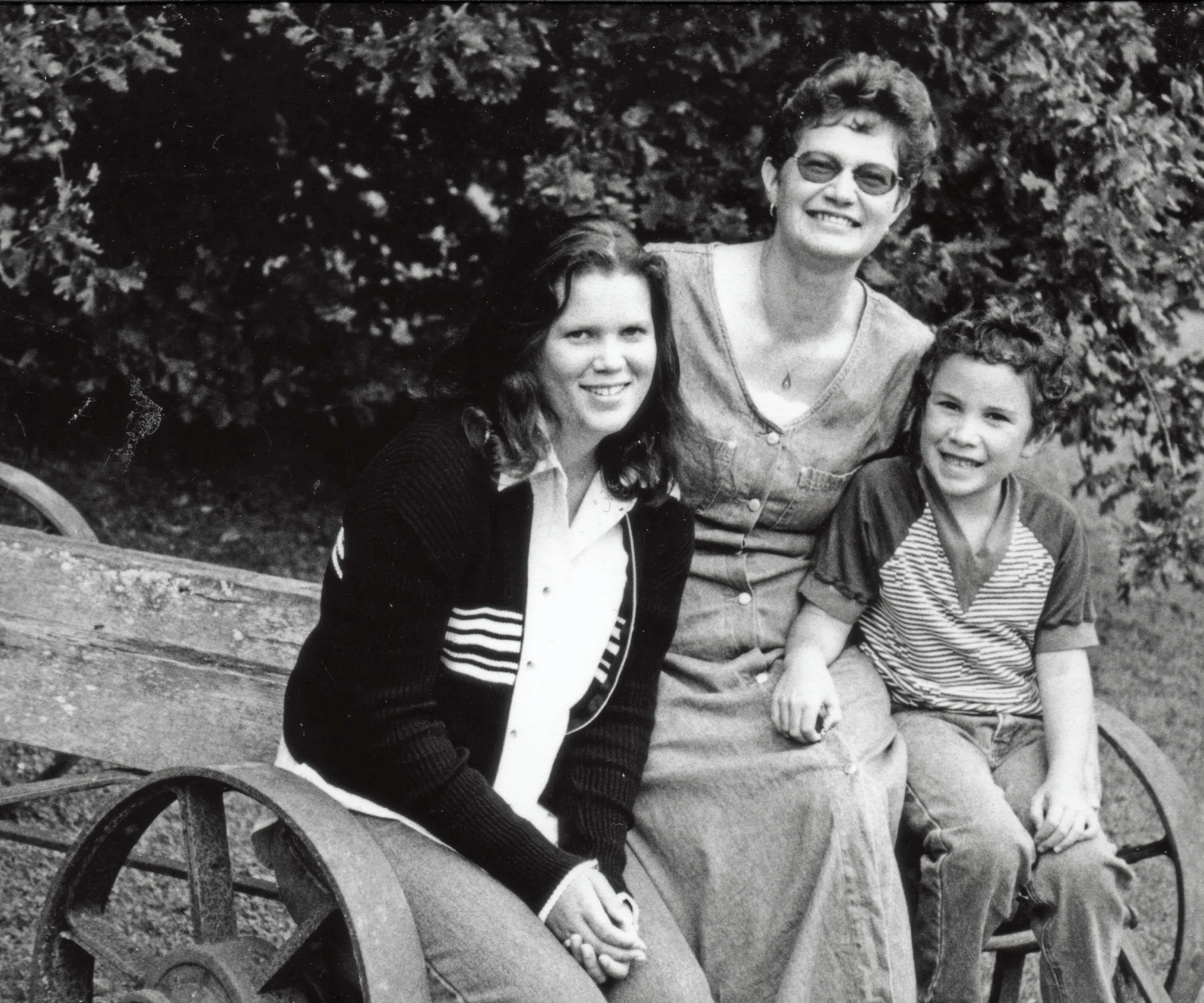 Three people, two adults and one child, sitting outdoors on a wooden bench, smiling at the camera.