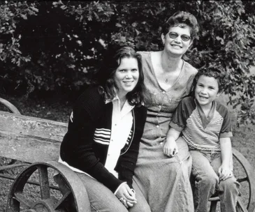 Three people, two adults and one child, sitting outdoors on a wooden bench, smiling at the camera.