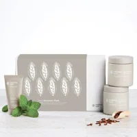 Skincare product set with two jars, a tube, mint leaves, cacao pieces, and a box featuring leaf patterns on a white surface.