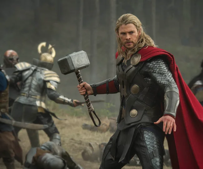 Thor in armor and red cape holds a hammer amidst a battle scene in a forest setting.