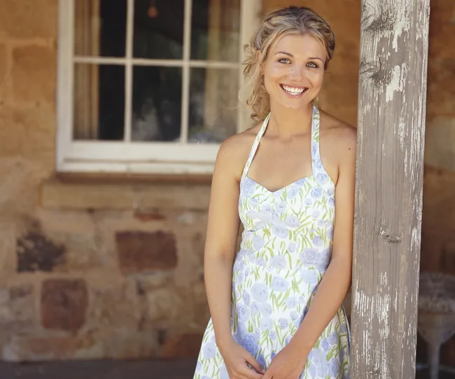 Woman in a floral dress smiles while leaning against a wooden post outside a rustic stone house.