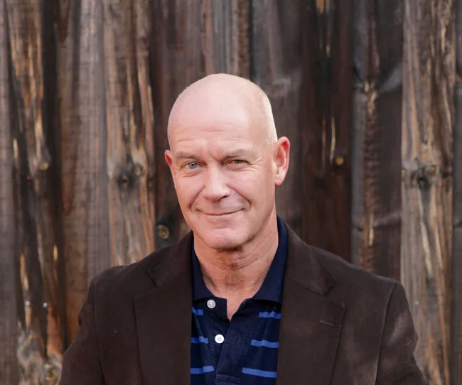 A man with a bald head and smiling expression standing in front of a wooden background, wearing a dark jacket and striped shirt.