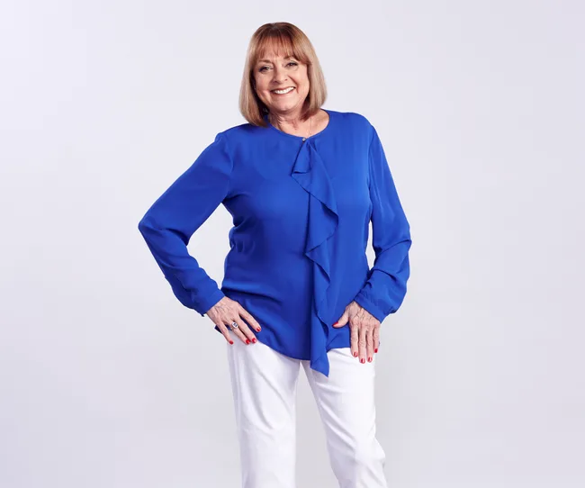 A woman in a blue blouse and white pants smiles, standing with one hand on her hip against a plain background.