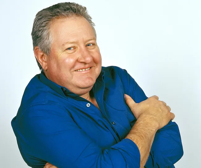 A person with short hair smiling, wearing a blue shirt, arms crossed against a plain background.