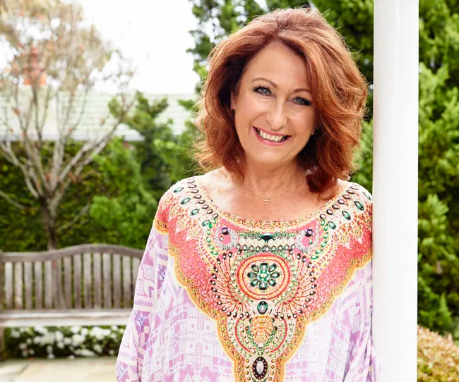 A woman with auburn hair smiles, wearing a colorful, patterned top, standing in a garden setting.