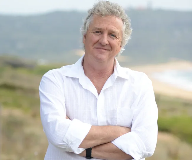 Man in a white shirt with folded arms standing outdoors with a grassy, blurred background.