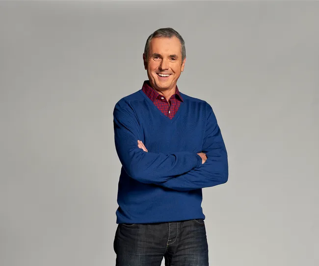 Man in a blue sweater and red checkered shirt, smiling with arms crossed against a plain background.