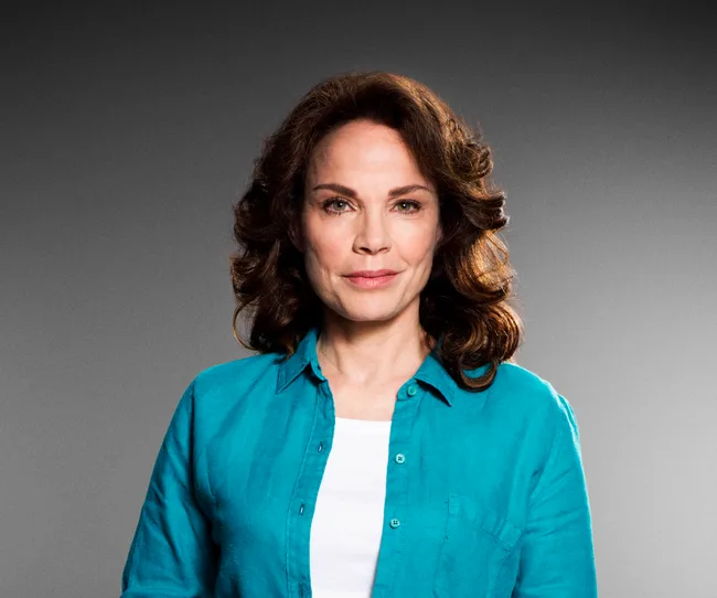 Woman with brown hair in a turquoise shirt, standing against a gray background.