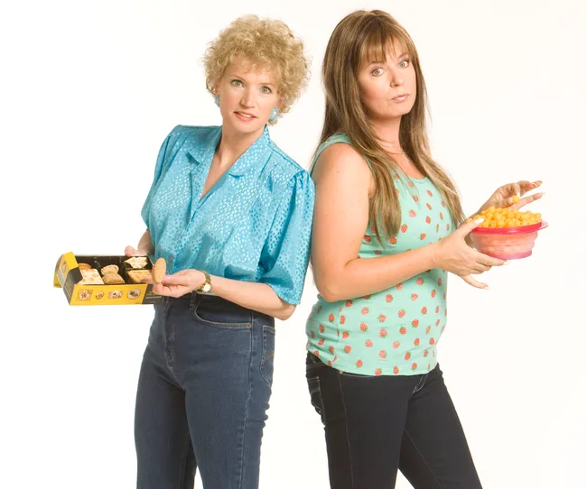 Two women standing back-to-back; one holds cookies, the other holds a bowl of cheese puffs, both in casual attire.