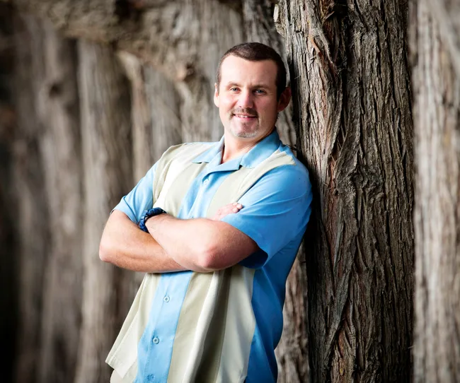 Man in blue shirt leans against a tree, arms crossed, smiling warmly.