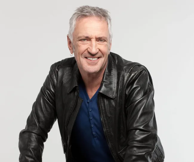 Man with gray hair in a black leather jacket smiling at the camera against a plain background.