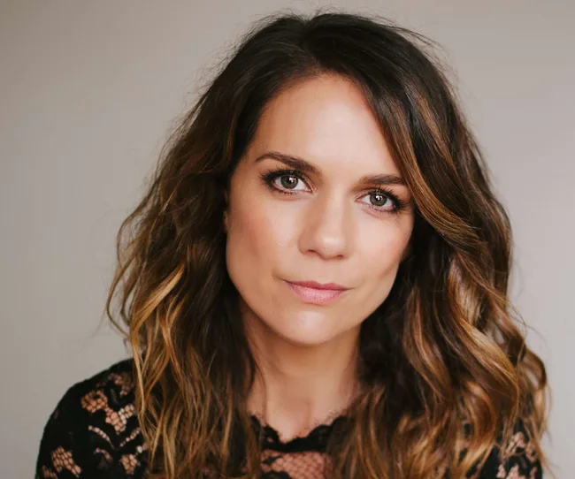 I don’t know who this is, but it's an image of a woman with wavy brown hair wearing a black lace top, facing the camera.