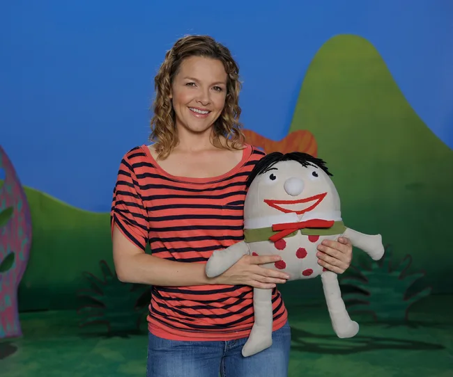 Woman holding a plush toy on a colorful set with greenery in the background.