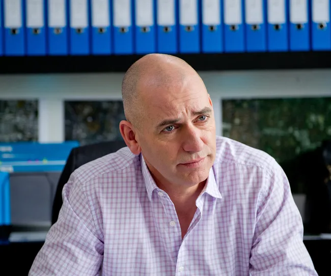 Bald man in a checked shirt sitting at a desk with blue binders in the background.