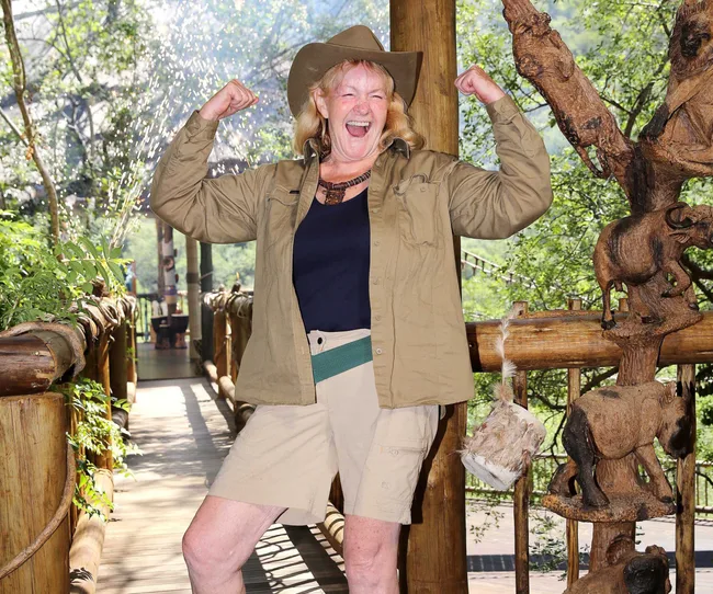 A woman in safari attire, flexing arms and smiling energetically, stands on a wooden bridge in a lush, leafy setting.