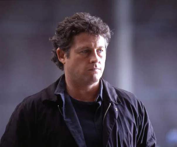 A man with curly hair wearing a dark jacket stands with a serious expression against a blurred background.