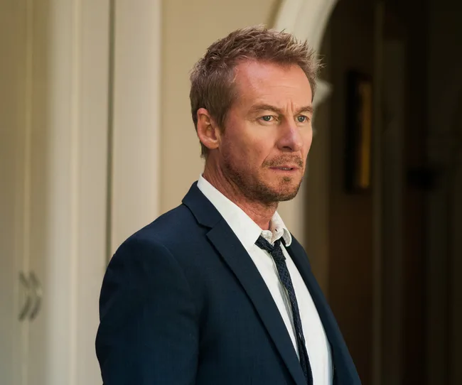 Man in a suit with messy hair looking thoughtful indoors.