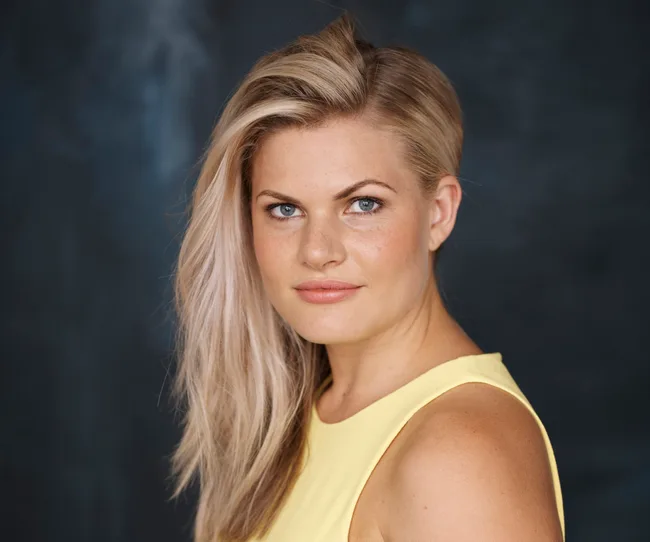 Blonde woman in a yellow top, looking confidently at the camera against a dark background.