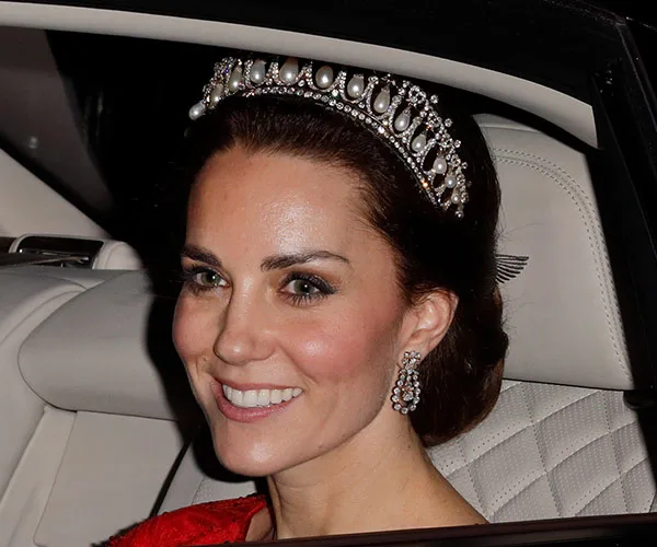A woman wearing a pearl and diamond tiara, smiling while seated in a car, dressed elegantly.