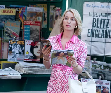 Woman in pink dress, reading a magazine at a newsstand, surrounded by newspapers and magazines.