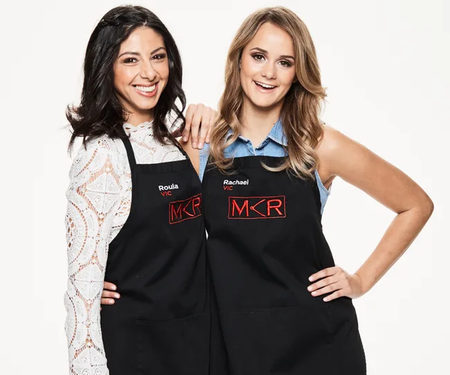 Two women in "MKR" aprons smiling, one with long dark hair and a white lace top, the other with lighter hair and a denim top.