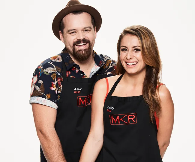 Two people wearing "MKR" aprons, smiling. One wears a hat and printed shirt, the other has long brown hair.